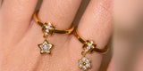 Two gold charm rings on a hand, one with a swinging crystal star and one with a crystal flower