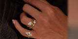 A hand against a dark ribbed sweater wearing a gold pear crystal ring and a kite-shaped crystal ring