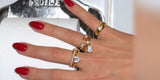 A hand with red nails wearing Oomiay gold rings on a bathroom sink
